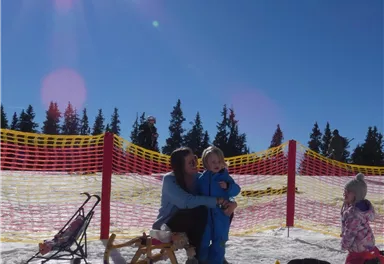 A mother is sitting in the snow with her child and hugging it. In the background, there are trees and a sunny sky.