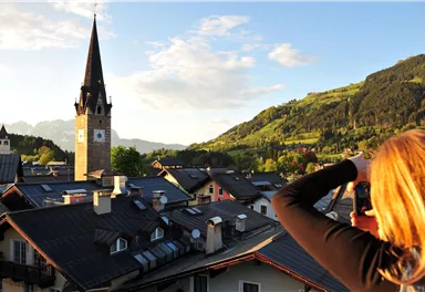 Eine Frau fotografiert die Aussicht auf ein malerisches Dorf mit einem hohen Kirchturm. Im Hintergrund sind grüne Hügel und ein klarer Himmel zu sehen.
