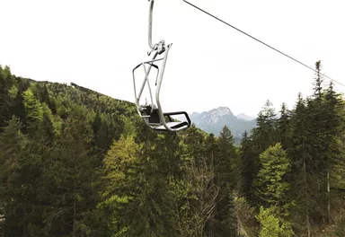 Ein Skilift schwebt über einen Wald mit dichten, grünen Bäumen. Im Hintergrund sind Berge zu sehen.