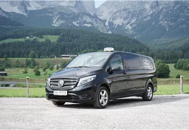 A black van from Mercedes-Benz is parked on a gravel-like area in front of a picturesque mountain landscape. In the background, green meadows and tall mountains are visible.