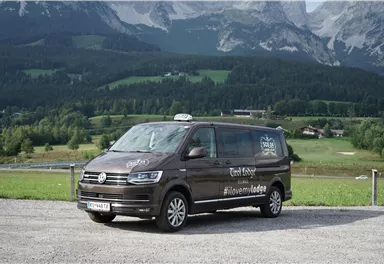 A brown Volkswagen taxi van is parked in a lot with a picturesque mountain landscape in the background. The surroundings are green and hilly under a clear sky.