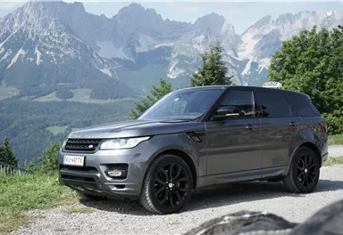 An elegant SUV stands on a gravel path in front of an impressive mountain landscape. The picturesque mountains and the clear sky give the scene a majestic atmosphere.
