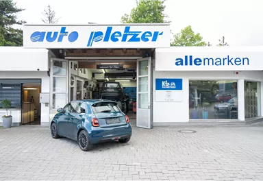 Eine Werkstatt mit dem Namen „auto pletzer“ und einem modernen Auto im Vordergrund. Im Hintergrund sind weitere Fahrzeuge und die Aufschrift „alle marken“ sichtbar.