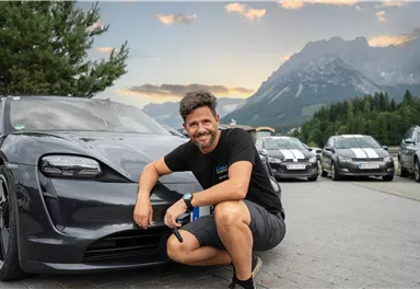 Ein Mann posiert lächelnd neben einem modernen Auto in einer bergigen Landschaft. Im Hintergrund sind weitere Autos und ein schöner Himmel zu sehen.