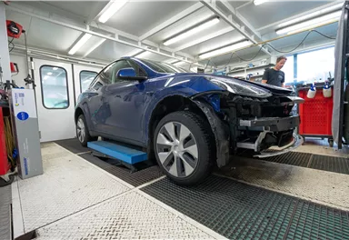 Ein blauer Wagen steht in einer Werkstatt auf einem Hebebühne. Im Hintergrund arbeitet eine Person an einem Auto.