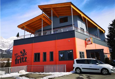 Ein modernes Gebäude mit einem orangefarbenen und grauen Design. Es hat einen Balkon und befindet sich in einer bergigen Umgebung.
