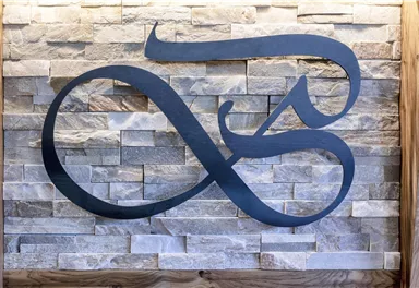 Ein stilvolles Logo aus schwarzem Metall steht vor einer Wand aus Naturstein. Das Design ist modern und ansprechend.