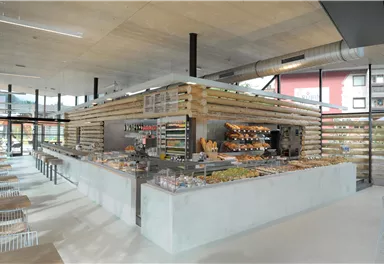 A modern café with bright, open spaces and a large counter that offers various food items. The decor is made of wood and provides an inviting atmosphere.
