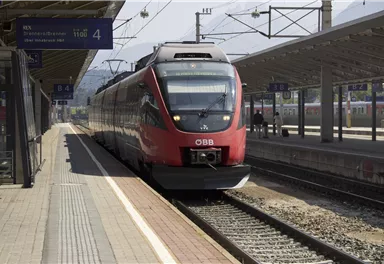 A red train from ÖBB is standing at a station. The tracks are clearly visible and the platform is empty.
