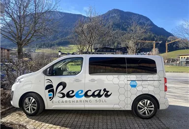 An e-carsharing vehicle of the brand Beecar is standing at the edge of a road. In the background, mountains and a clear sky are visible.