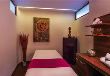 Ein entspannender Behandlungsraum mit einem Massagetisch und sanfter Beleuchtung. Im Raum befinden sich dekorative Elemente und Handtücher, die für eine ruhige Atmosphäre sorgen.