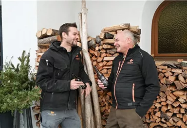 Zwei Männer stehen lächelnd vor einem Holzstapel. Sie halten Flaschen in der Hand und scheinen sich bestens zu unterhalten.