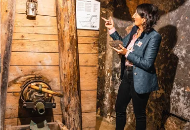 Eine Frau steht in einem Bergwerk und erklärt eine Informationstafel. Um sie herum sind Holzelemente und Bergbaugeräte sichtbar.