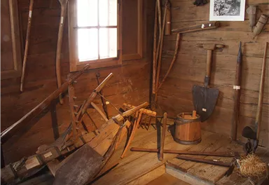 An old workshop with various agricultural tools. The walls are made of wood and a window lets light in.