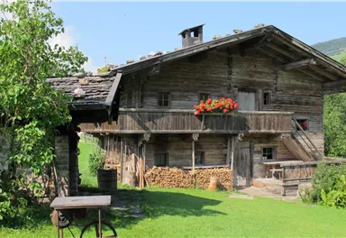 A traditional wooden house with a balcony and red flowers. The garden is green and well-maintained, surrounded by mountains.