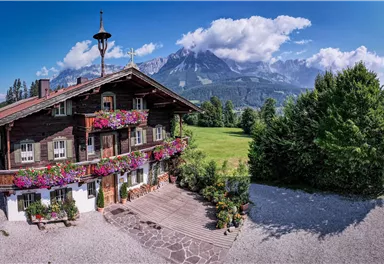 Ein malerisches Bauernhaus mit bunten Blumen und einer idyllischen Umgebung. Im Hintergrund erheben sich majestätische Berge unter einem blauen Himmel.