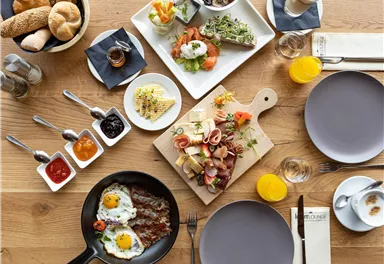 A rich breakfast buffet with various dishes. There are eggs, cold cuts, bread, fruit, and different sauces on a wooden board.