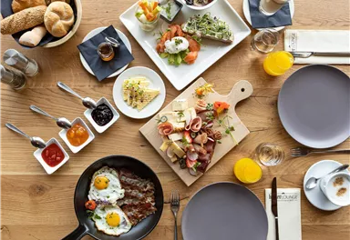 A rich breakfast buffet with various dishes, including eggs, sausages, and fresh vegetables. There is also bread, jam, and drinks.