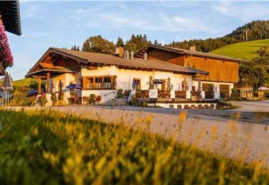 A traditional building in a rural style, surrounded by meadows and forests. The terrace is inviting with tables and sun umbrellas.