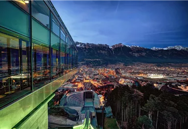 A modern building view with large windows overlooking a illuminated cityscape and the surrounding mountains. It is evening, and the sky is tinted in gentle shades of blue and violet.