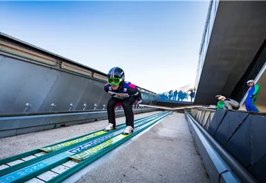 A ski jumper is ready on the ramp and is preparing for the jump. In the background, spectators and the impressive architecture of the jump facility can be seen.