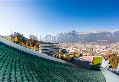 A view of the city with mountains in the background and clear blue sky. In the foreground, the ski jumping facilities and the surrounding landscape can be seen.
