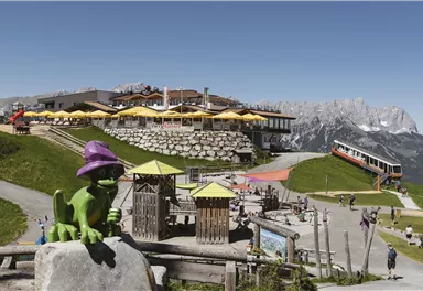A mountain landscape with a restaurant and sunny terraces. In the foreground stands a green figure with a purple hat and playground equipment for children.