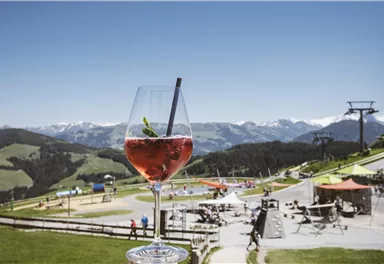 A refreshing drink in a glass is in the foreground. In the background, a beautiful mountain landscape and a recreational area can be seen.