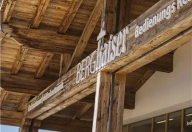 A rustic wooden building with the sign "Mountain Cheese Factory" and the inscription "Service Restaurant". In the foreground, colorful flowers can be seen.