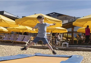 A boy joyfully jumps on a trampoline. In the background, there are sunny loungers and sun umbrellas.