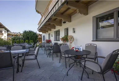 A cozy terrace with tables and chairs. In the background, plants and a clear sky can be seen.