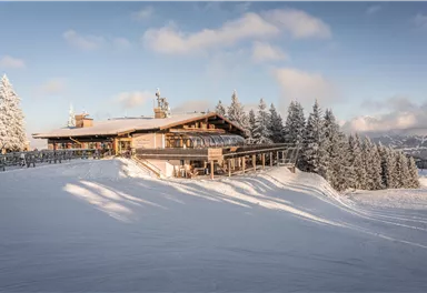Ein großes Restaurant in den Bergen, umgeben von schneebedeckten Tannen. Der Himmel ist klar und sonnig, ideal für Wintersport.