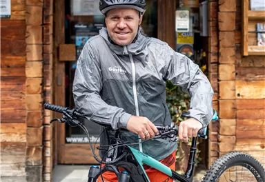 A man in a gray rain jacket stands next to a mountain bike. In the background, a wooden cabin is visible.