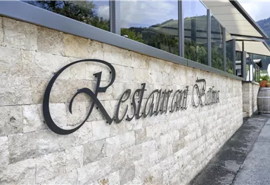 Ein elegantes Restaurant mit dem Schriftzug "Restaurant Bellevue" an einer Steinwand. Im Hintergrund sind grüne Hügel und ein klarer Himmel sichtbar.