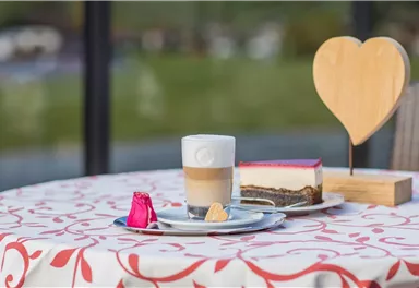 Ein schöner Tisch mit einem Latte Macchiato, einem Stück Kuchen und einer herzförmigen Holzdekoration. Der Tisch ist mit einer eleganten Tischdecke gedeckt und bietet eine angenehme Atmosphäre.