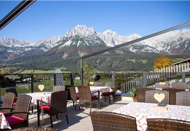 Eine gemütliche Terrasse mit Blick auf hohe, schneebedeckte Berge. Die Tische sind mit roten Sitzkissen ausgestattet und die Umgebung ist grün und einladend.