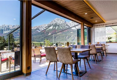 Ein modernes Restaurant mit Holzdekor und Tischen ist zu sehen. Durch große Fenster bietet sich ein spektakulärer Blick auf die Berge.