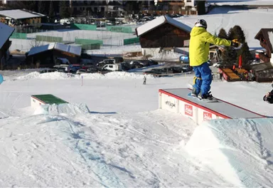 A snowboarder is riding over a rail in the snow. In the background, snow-covered cabins and a ski slope can be seen.