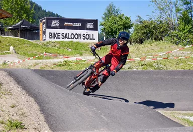 A mountain biker is riding on a winding track. In the background, the sign "Bike Saloon Söll" is visible.