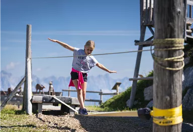 A girl is balancing on a rope in the mountains. In the background, a beautiful landscape and a wooden structure are visible.