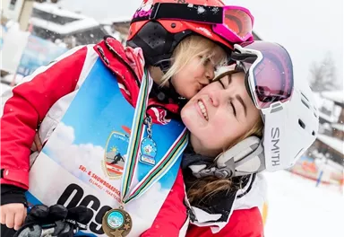 A happy girl kisses a skier with a medal. Both are wearing winter ski clothes and are smiling on a snowy slope.
