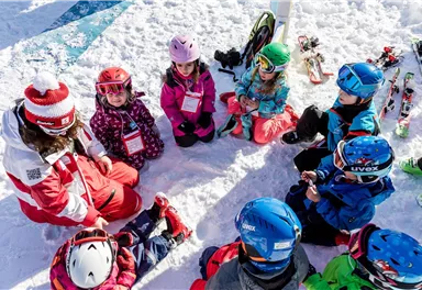 A group of children is sitting in the snow and listening to a ski instructor. Surrounding them are skis, and it seems to be sunny.