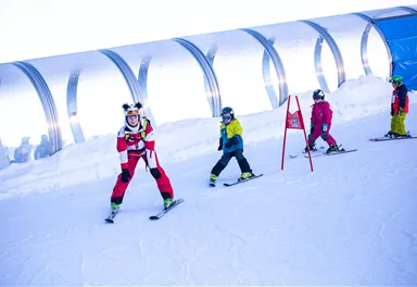 A cheerful ski school on the slope with several children in colorful ski gear. In the background, a large, bright ski hall can be seen.