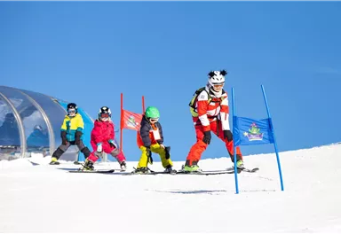 A group of children learning to ski stands on a snow-covered slope. In the background, colorful flags and a blue sky can be seen.