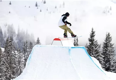 A snowboarder jumps over a stylized snow park element in a snowy landscape. In the background, there are visible pine trees and mountain peaks.