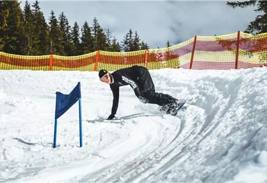 A snowboarder is riding through the snow and has taken a turn. In the background, there are snow-covered trees and a yellow barrier visible.