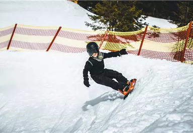 A boy is snowboarding in the snow. Safety nets are visible in the background.