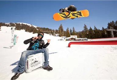 A snowboarder jumps high in the air over a snow slope. In the foreground, a man is sitting and watching the action.