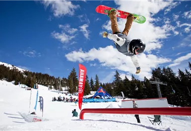 A snowboarder is performing a trick in the air. In the background, snow-covered mountains and spectators can be seen.