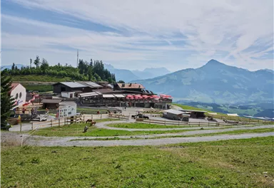 Eine malerische Berglandschaft mit einem Gasthof und roten Sonnenschir men. Die Umgebung ist grün mit sanften Hügeln und Bergen im Hintergrund.
